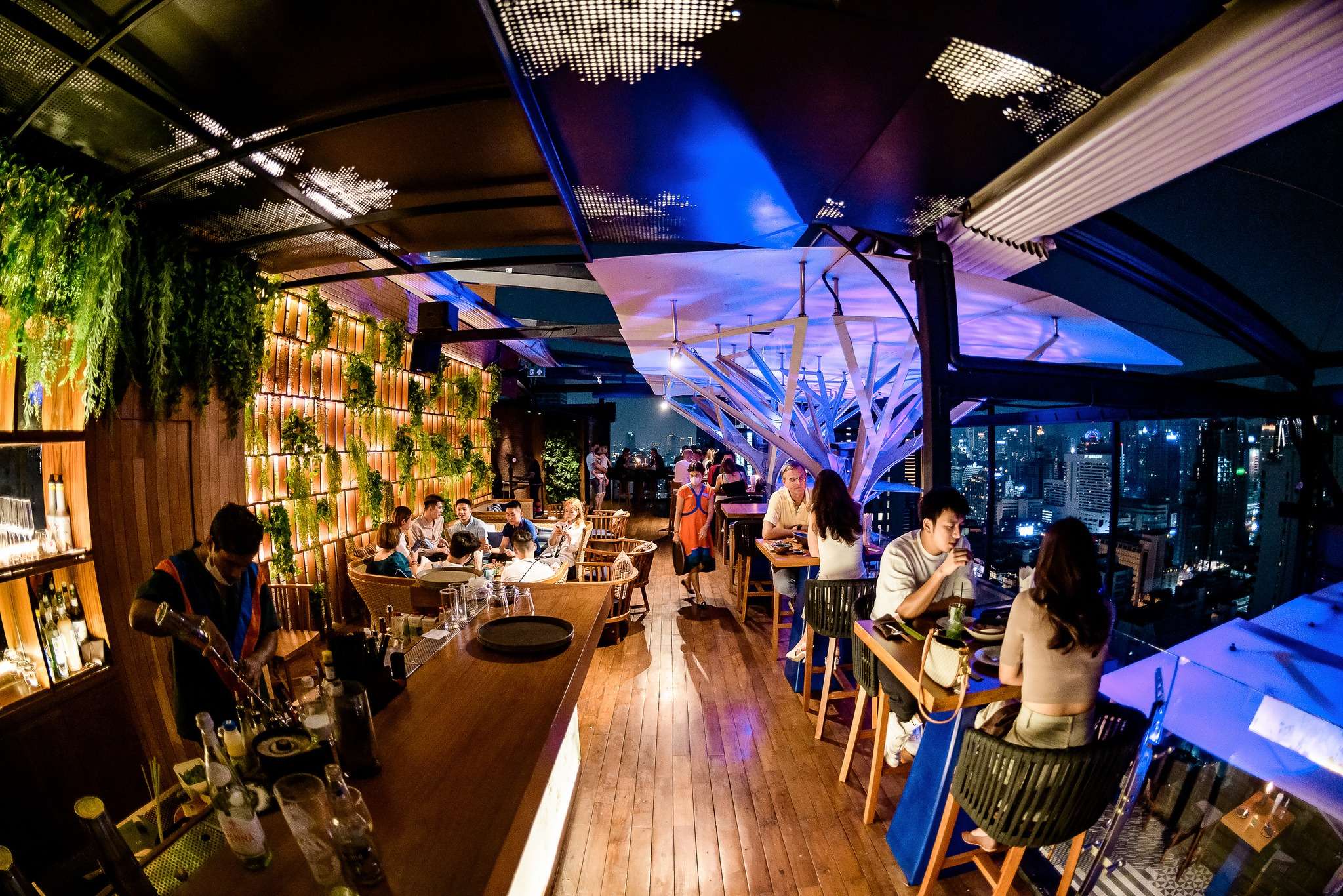 Above Eleven's rooftop interior — the sculptural white tree, living-green walls and warm wood over the Bangkok skyline